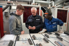 KI generiert: Drei Personen in Arbeitskleidung stehen an einem Tisch mit Metallstücken und scheinen sich über eine technische Aufgabe zu unterhalten.