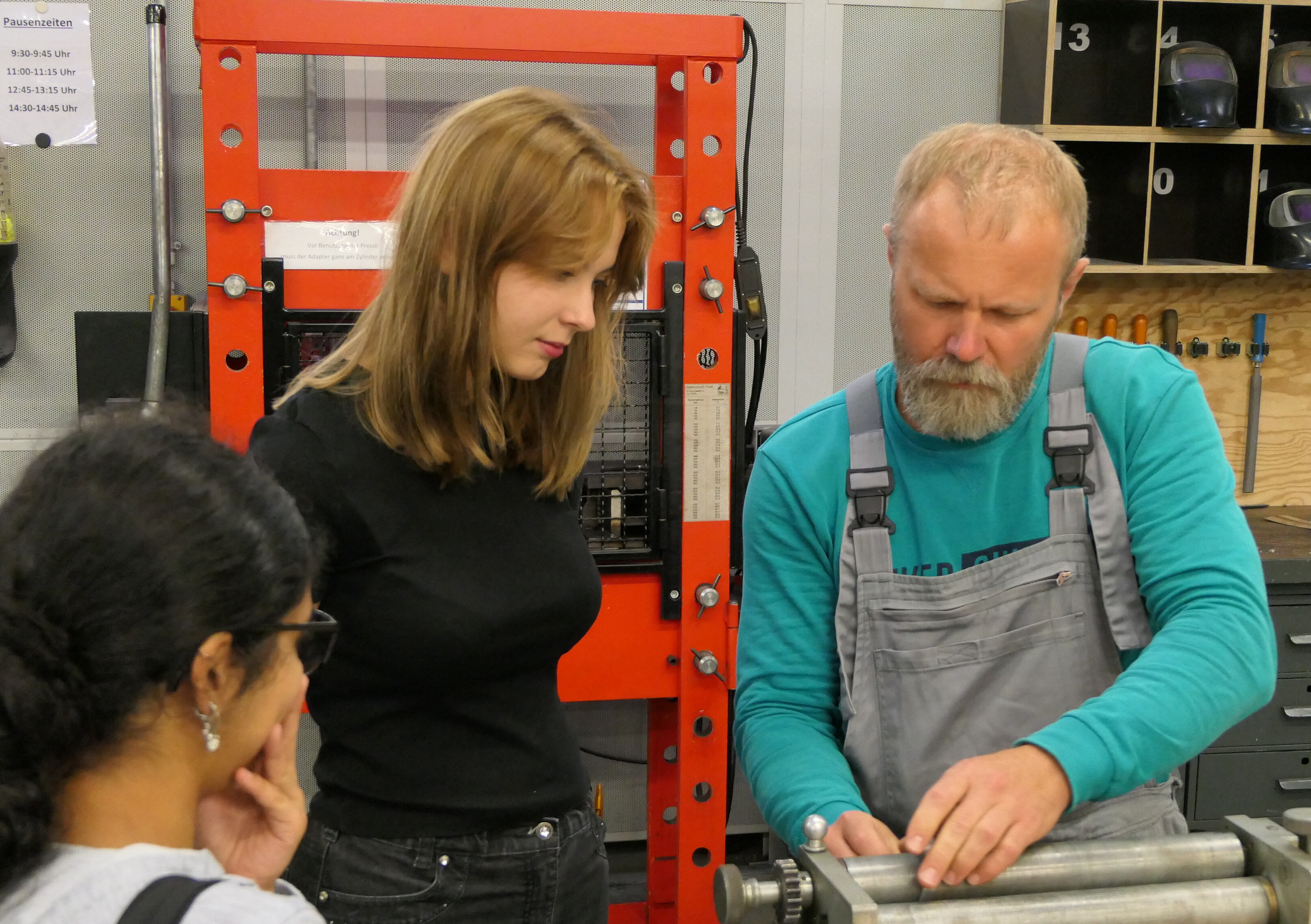 KI generiert: Ein Mann in Arbeitskleidung zeigt zwei Frauen in einem technischen Umfeld ein Gerät oder eine Maschine.