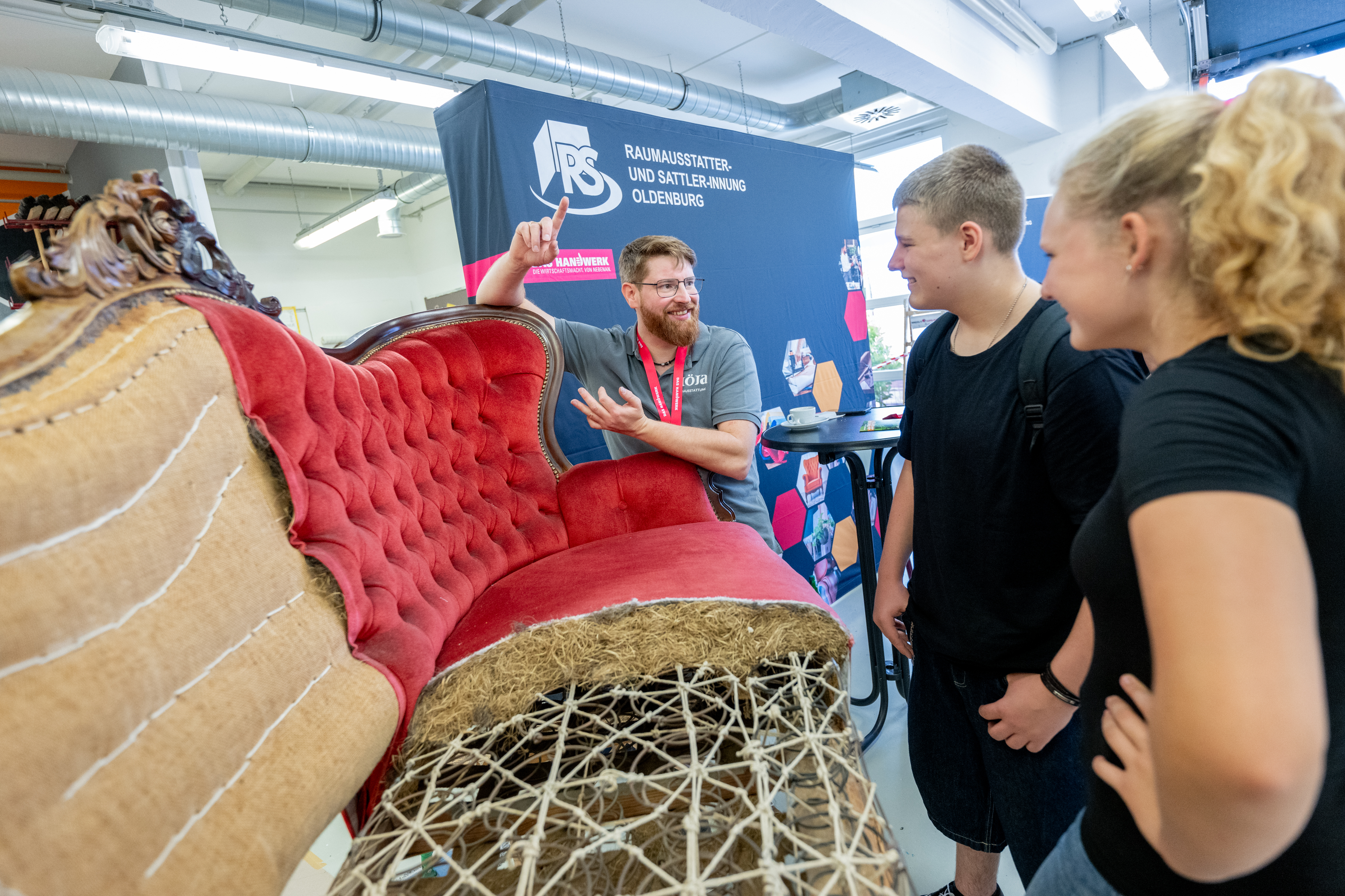 KI generiert: Das Bild zeigt eine Gruppe von Menschen, die an einem Stand auf einer Messe oder Ausstellung teilnehmen, wo sie Informationen über ein Produkt erhalten, das in einem Netz aus roter Folie verpackt ist.