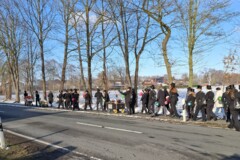 KI generiert: Eine Gruppe von Menschen mit Hüten zieht mit Handkarren auf einem winterlichen Bürgersteig entlang einer Straße.
