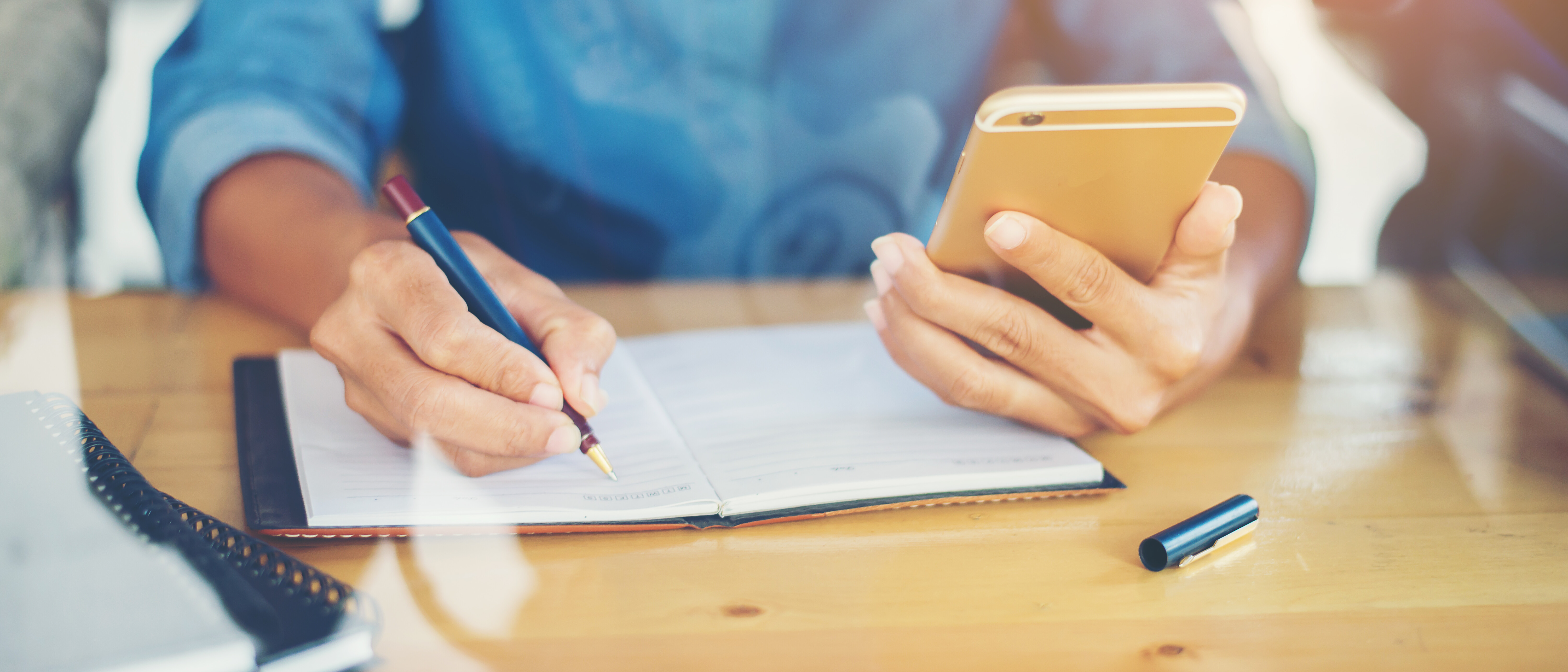KI generiert: Die Person schreibt in ein Notizbuch, während sie ein Smartphone hält.