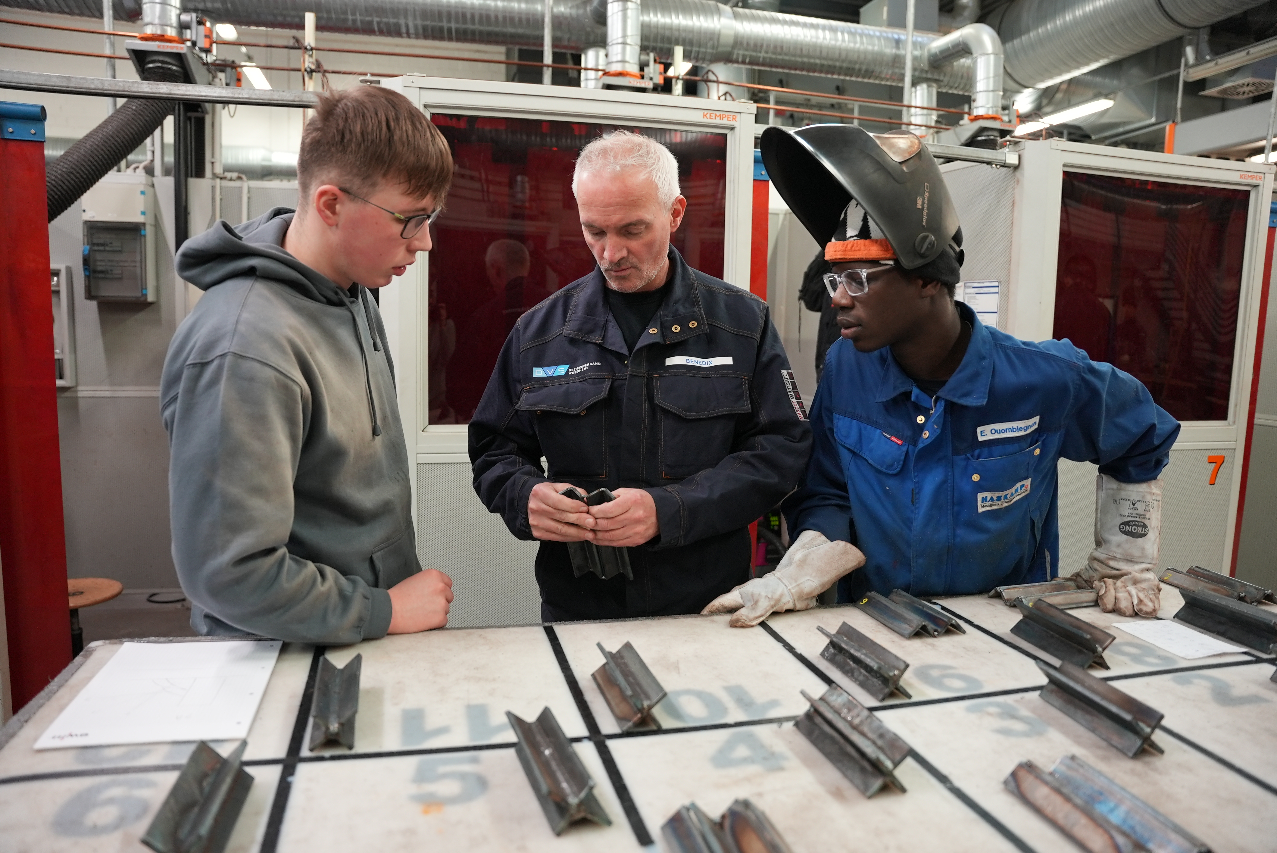 KI generiert: Drei Personen in Arbeitskleidung stehen an einem Tisch mit Metallstücken und scheinen sich über eine technische Aufgabe zu unterhalten.