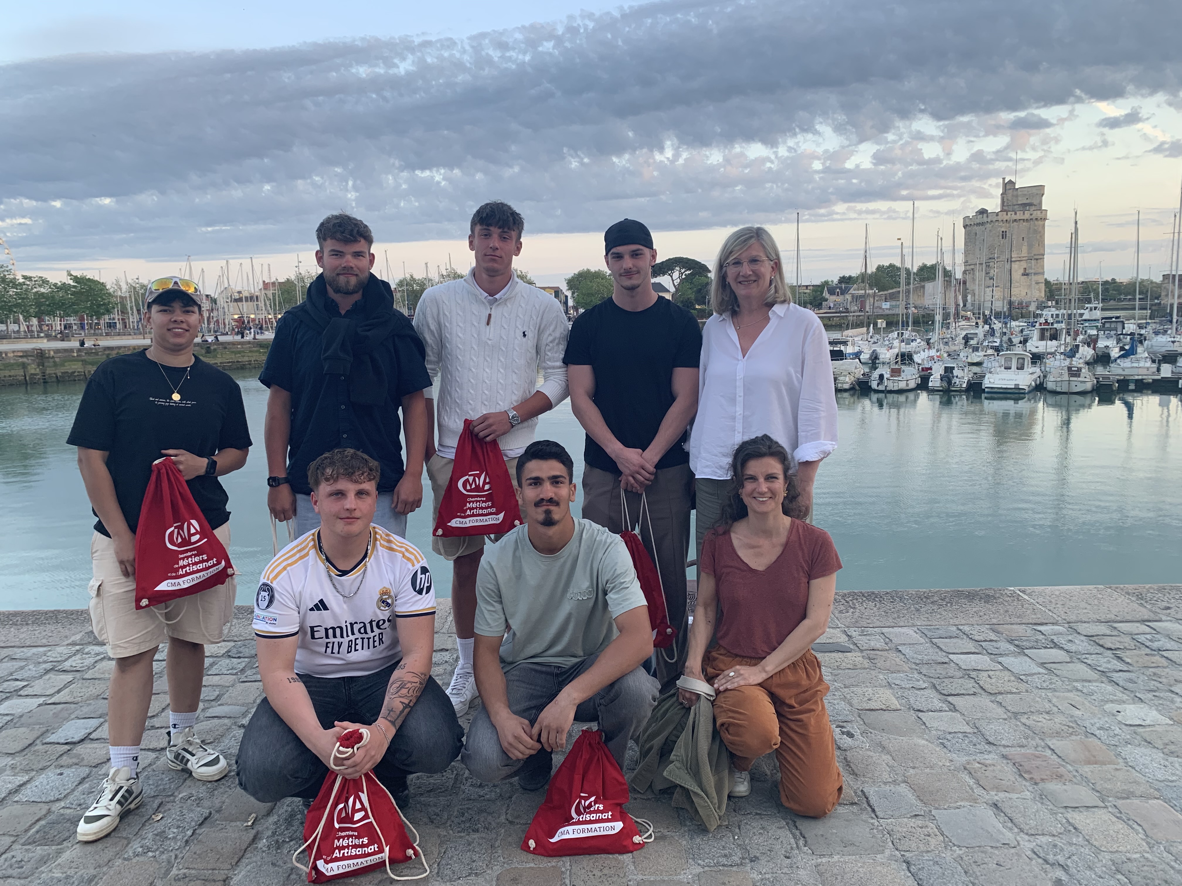 KI generiert: Das Bild zeigt eine Gruppe von sieben Personen, die auf einer Uferpromenade mit Blick auf einen Hafen und eine Stadt stehen und lächeln.