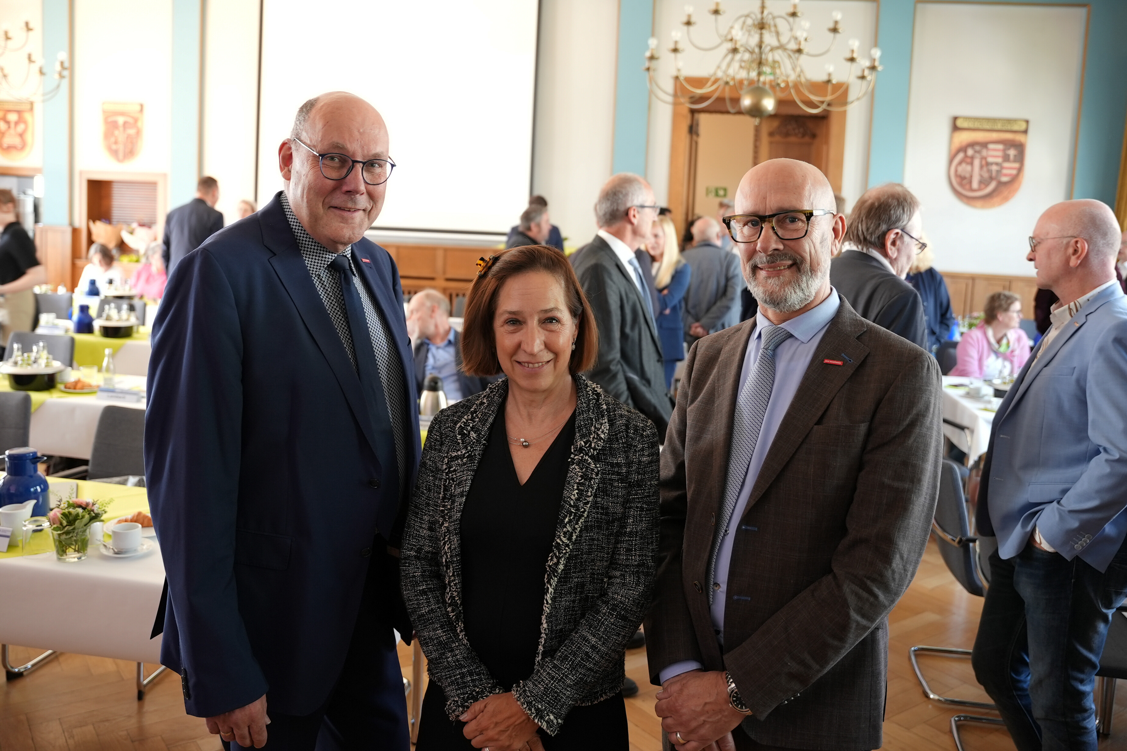 KI generiert: Das Bild zeigt drei Personen, die in einem formellen Veranstaltungsraum stehen.