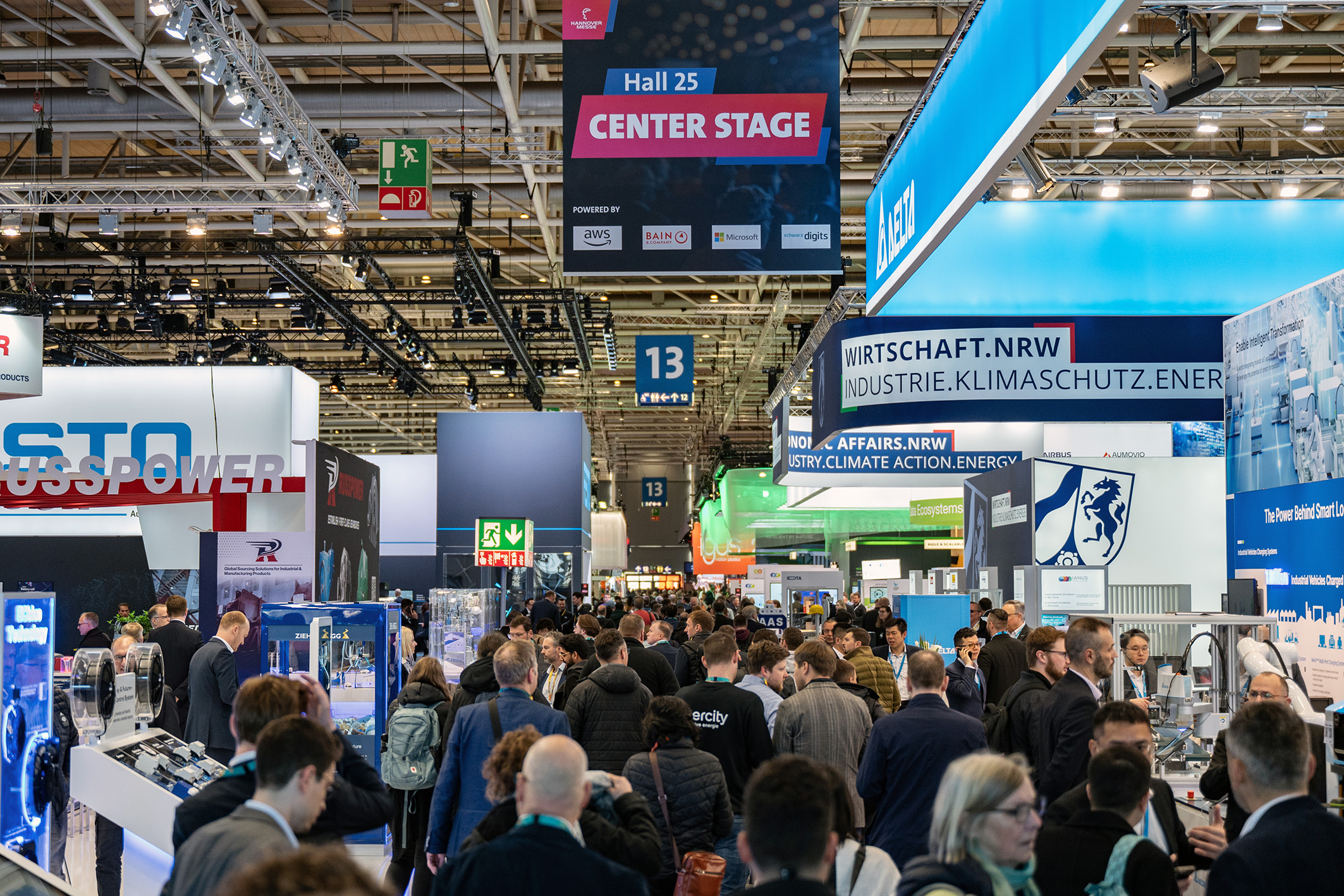 KI generiert: Das Bild zeigt eine belebte Messe mit vielen Besuchern, die sich an verschiedenen Ständen und Ausstellungen informieren, darunter ein großer Stand mit der Aufschrift "Center Stage" und ein Stand mit dem Logo "wirtschaft.nrw".