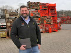 KI generiert: Ein Mann steht lächelnd vor einem Stapel von Metallkisten und hält ein Tablet in der Hand.