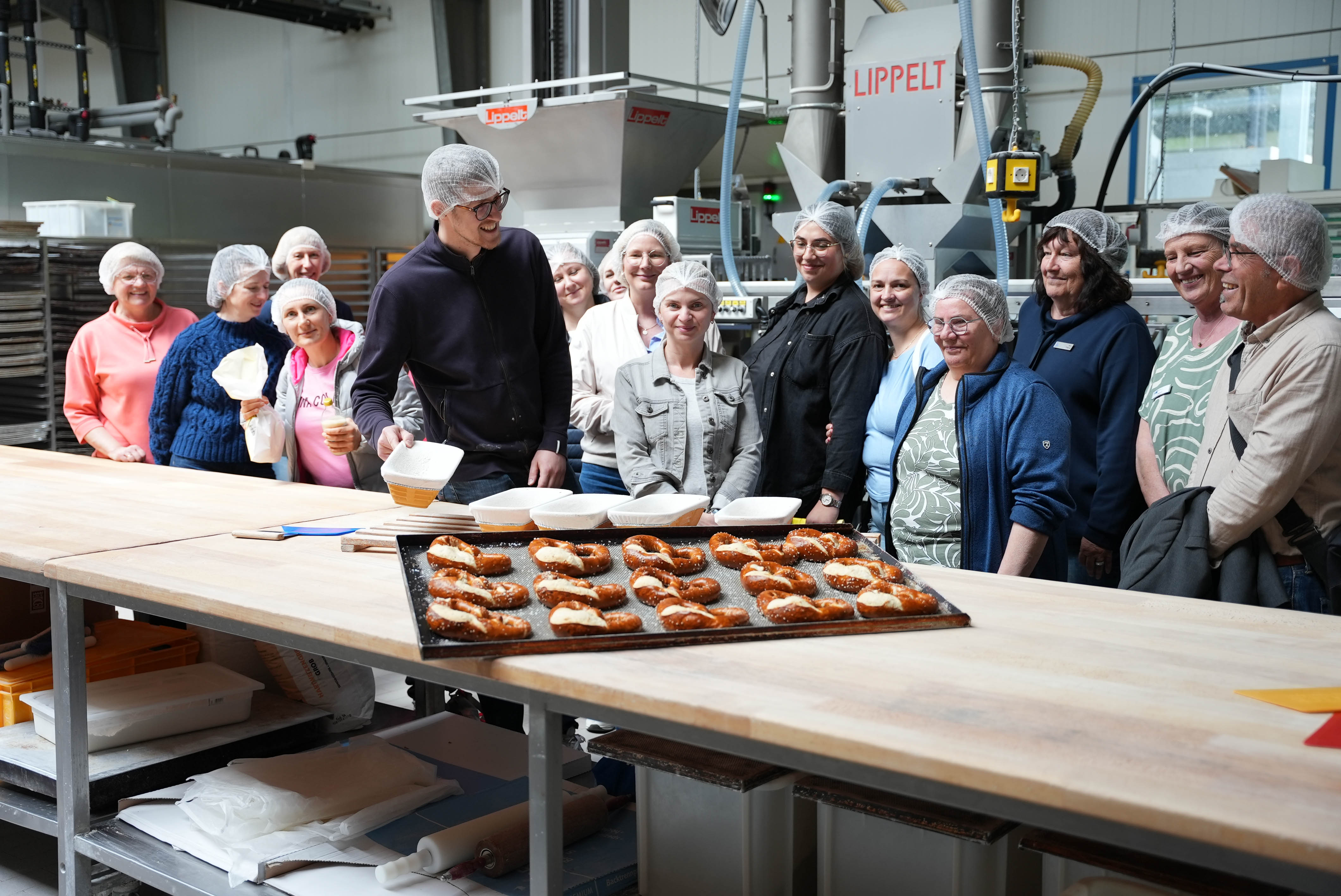 KI generiert: Eine Gruppe von Menschen mit Haarnetzen steht in einer Bäckerei um einen Tisch mit frisch gebackenen Brezeln.