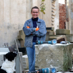 KI generiert: Eine Person steht lächelnd in einer Werkstattumgebung neben einem großen Steinblock mit Werkzeugen darauf, während ein Hund daneben sitzt.