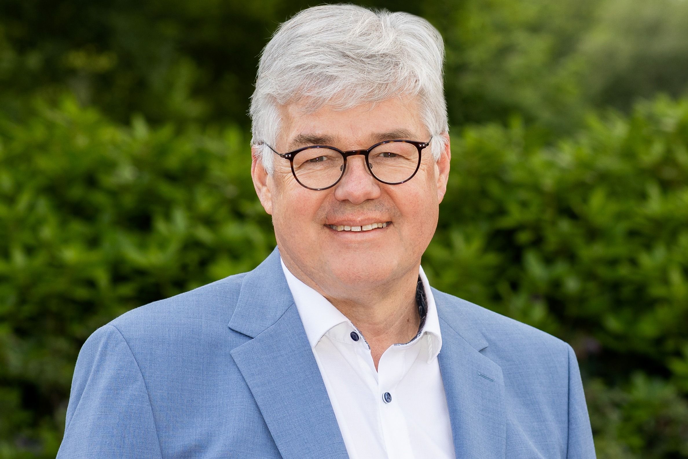KI generiert: Das Bild zeigt einen lächelnden Mann in einem blauen Anzug mit Brille vor einem grünen Hintergrund.