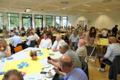 KI generiert: Das Bild zeigt eine große Gruppe von Menschen, die in einem Raum an verschiedenen Tischen bei einer Versammlung oder Veranstaltung sitzen.
