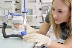 Zahngebiss im Vordergrund in Halterung eingespannt. Junge Frau mit Gummihandschuhen schaut auf Gebiss